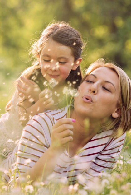 A woman and a girl blow dandelion seeds into the air.