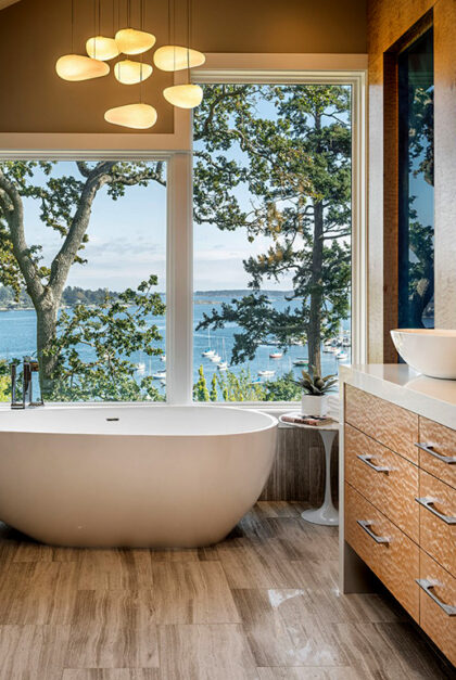 A photo of a bathroom, with a tub set against a window with a view of water, trees, and sailboats.